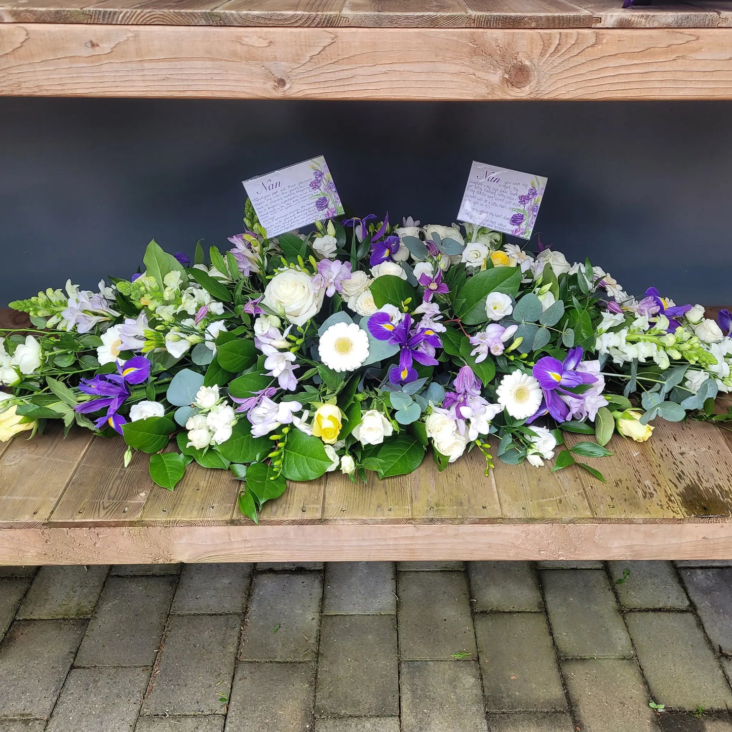 The Casket Spray Funeral Flowers by ReCreation Flowers features white and purple blooms, including roses and irises, arranged above a stone surface with two condolence cards nestled among the stems in a heartfelt tribute.