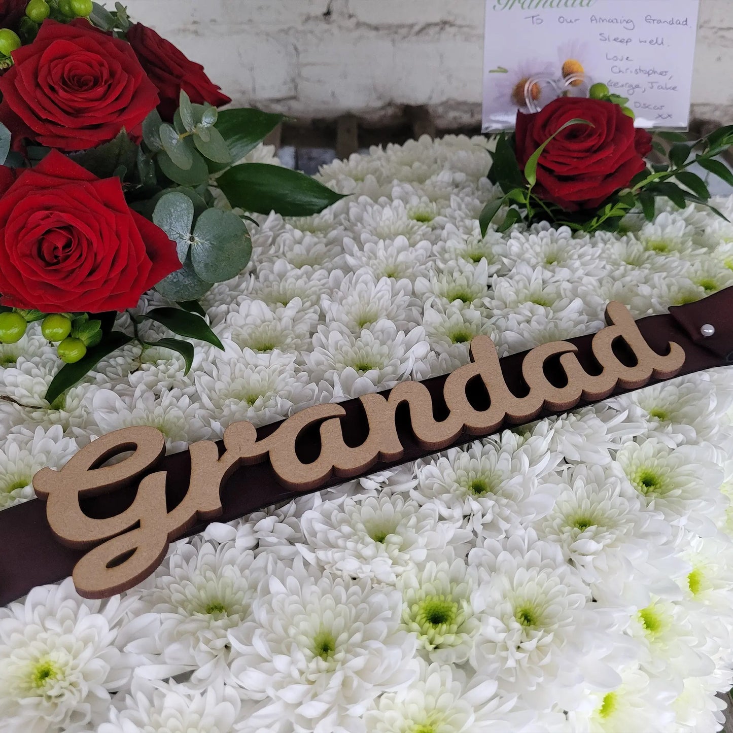 The Solid Heart Funeral Flowers by ReCreation Flowers features seasonal blooms, white chrysanthemums, red roses, greenery, a wooden "Grandad" sign on top, and a small card nestled among the arrangement.