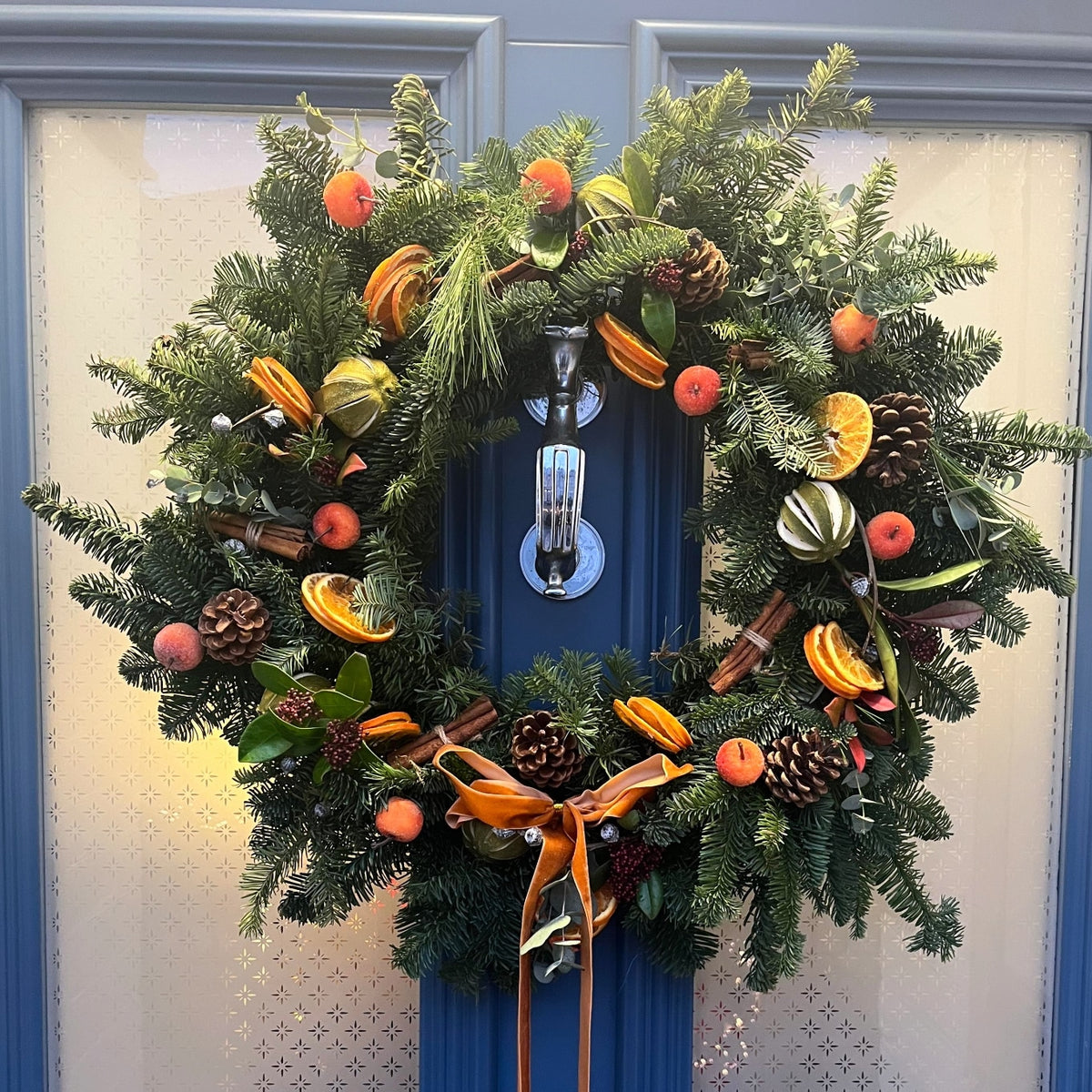 A festive wreath featuring pinecones, dried orange slices, small red ornaments, lush greenery, and a gold ribbon hangs on a blue doorâperfect inspiration after attending the Christmas Workshop @ GAS by ReCreation Flowers in Leigh on Sea.