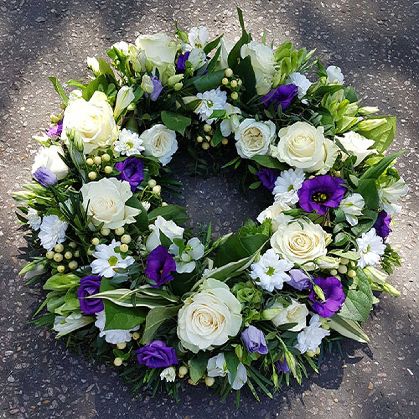 The Funeral Flower Wreath by ReCreation Flowers features white roses, purple blooms, small white petals, and lush greenery on a textured gray base—a tasteful arrangement ideal as funeral flowers.