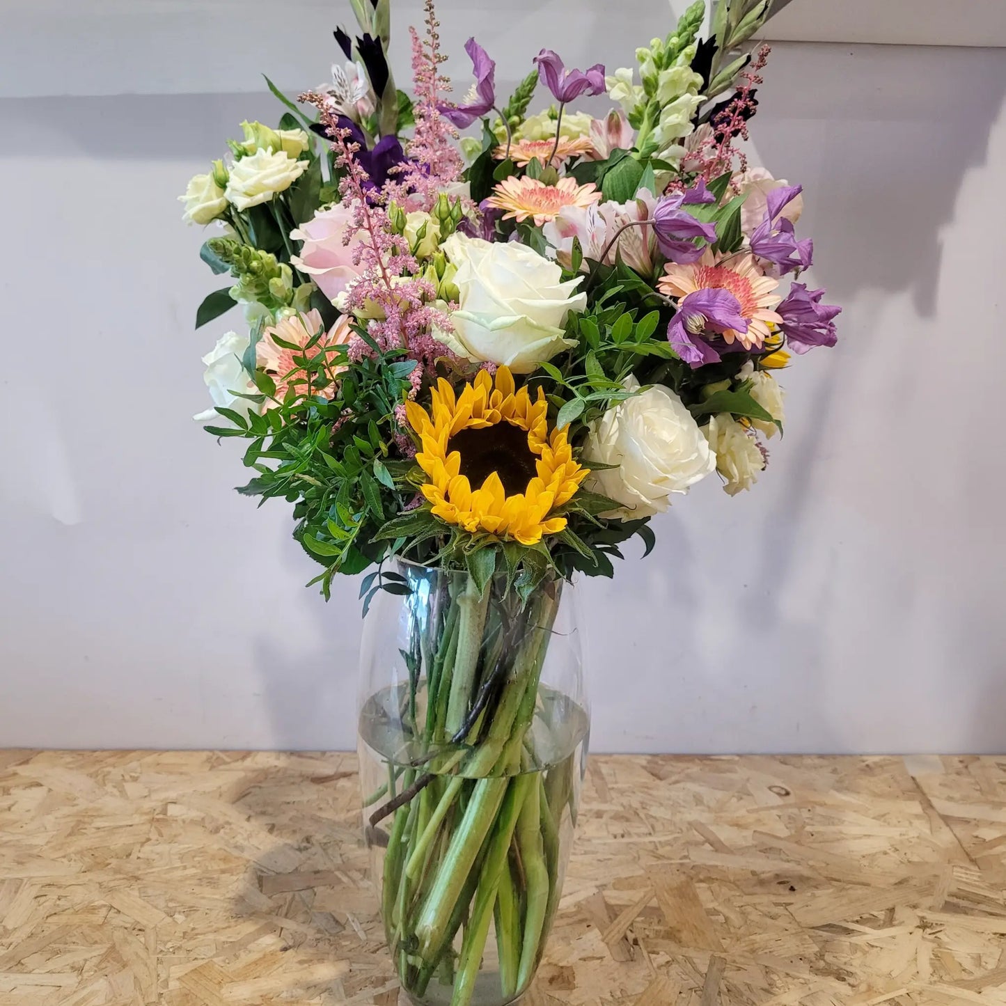 Scent With Love by ReCreation Flowers features a sunflower, white roses, pink and purple blooms, and greenery in a clear glass vase—lovingly arranged by your Leigh-on-Sea florist. Perfect for same-day delivery. Displayed on wood against a light wall.