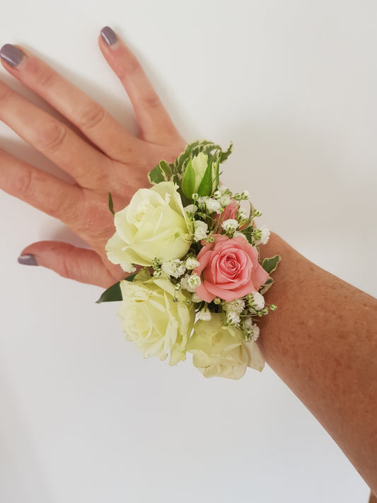 A hand with painted nails wears a floral wrist corsage made of white and pink roses, small white flowers, and green leaves against a plain white background.