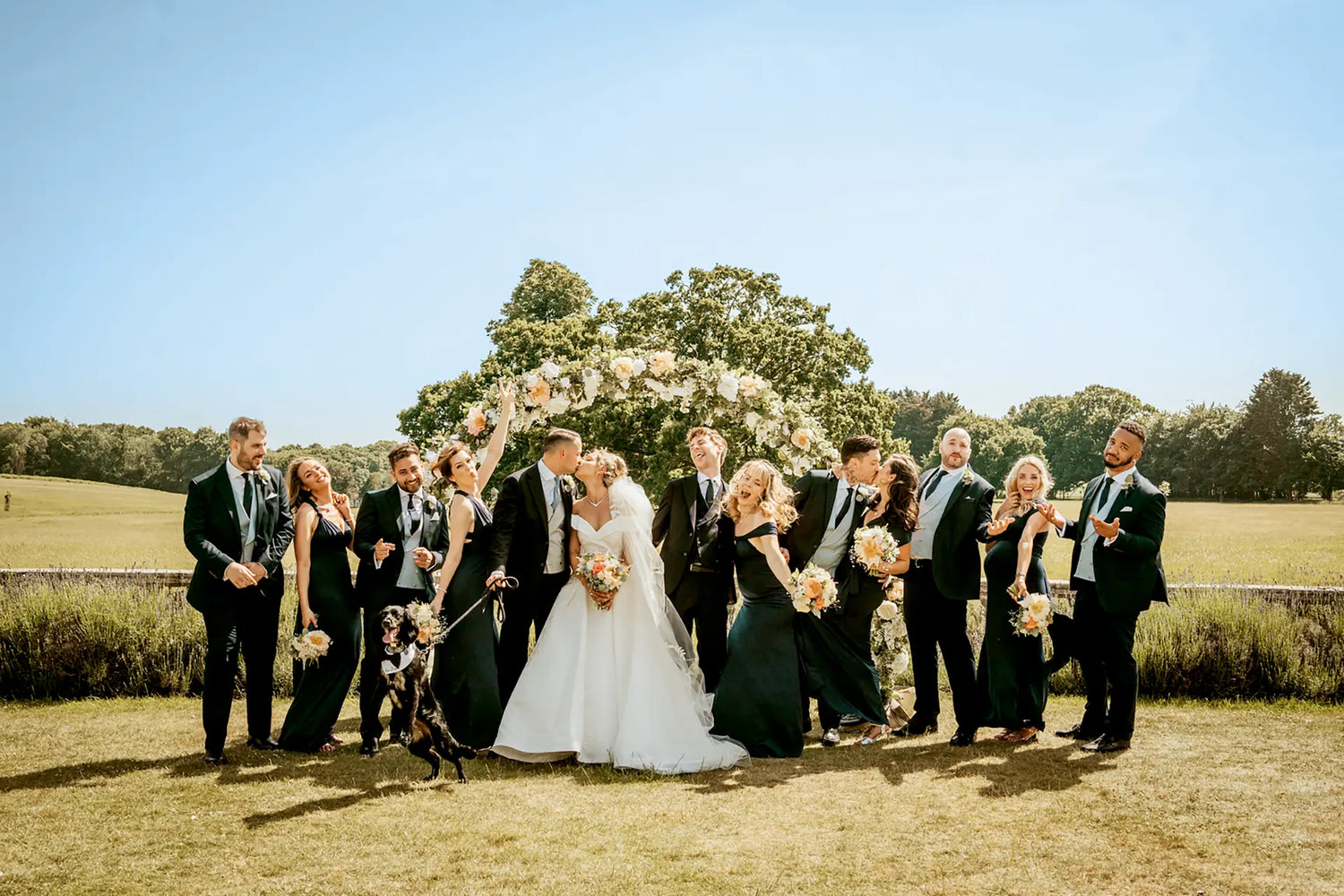 A wedding party poses outdoors; the bride and groom kiss under a floral arch, surrounded by joyful bridesmaids and groomsmen in dark dresses and suits, holding bouquets, with a dog in front.