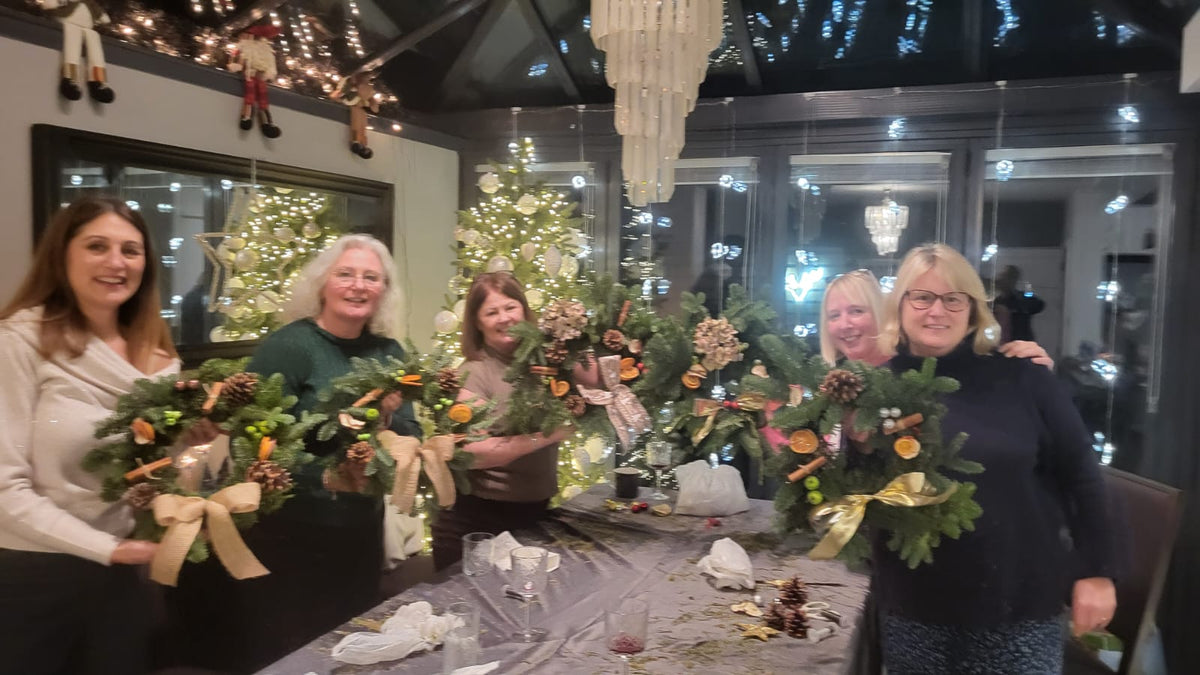 Four women smile and hold beautifully decorated Christmas wreaths in a festively lit room adorned with Christmas trees and twinkling lights. In front of them, a table filled with craft supplies highlights the creativity and joy experienced during the ReCreation Flowers Christmas Wreath Class in Leigh on Sea.