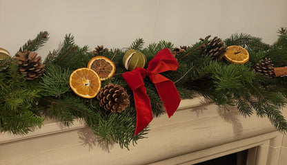 A beautiful garland featuring pine branches, pine cones, dried orange slices, and a red velvet bow sits on a white mantel—perfect inspiration if you’re joining the Christmas Garland Class by ReCreation Flowers or planning to craft your own fresh festive garland this season.