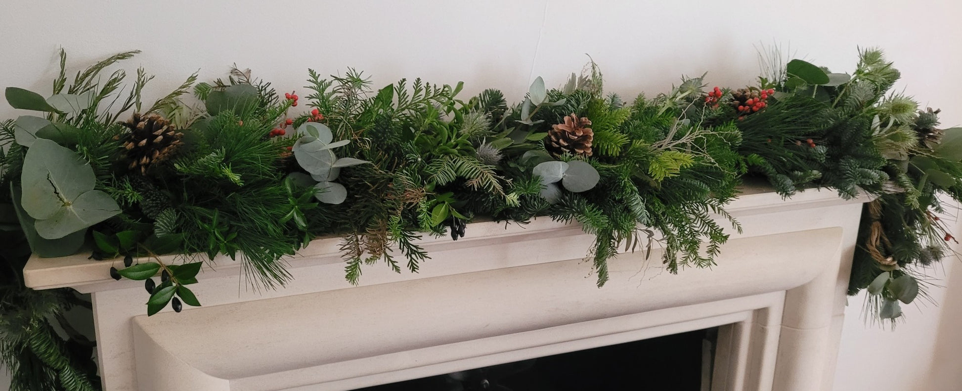 The Christmas Garland Class by ReCreation Flowers, featuring pinecones, red berries, and eucalyptus leaves, drapes festively over a white fireplace mantel.