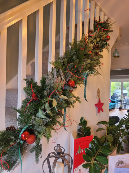 The Christmas Garland Class by ReCreation Flowers adorns the staircase railing with lush green garland, accented by red and teal ribbons, red baubles, and pinecones. Nearby, holiday cards, a star ornament, plants, and a lantern enhance the festive atmosphere.