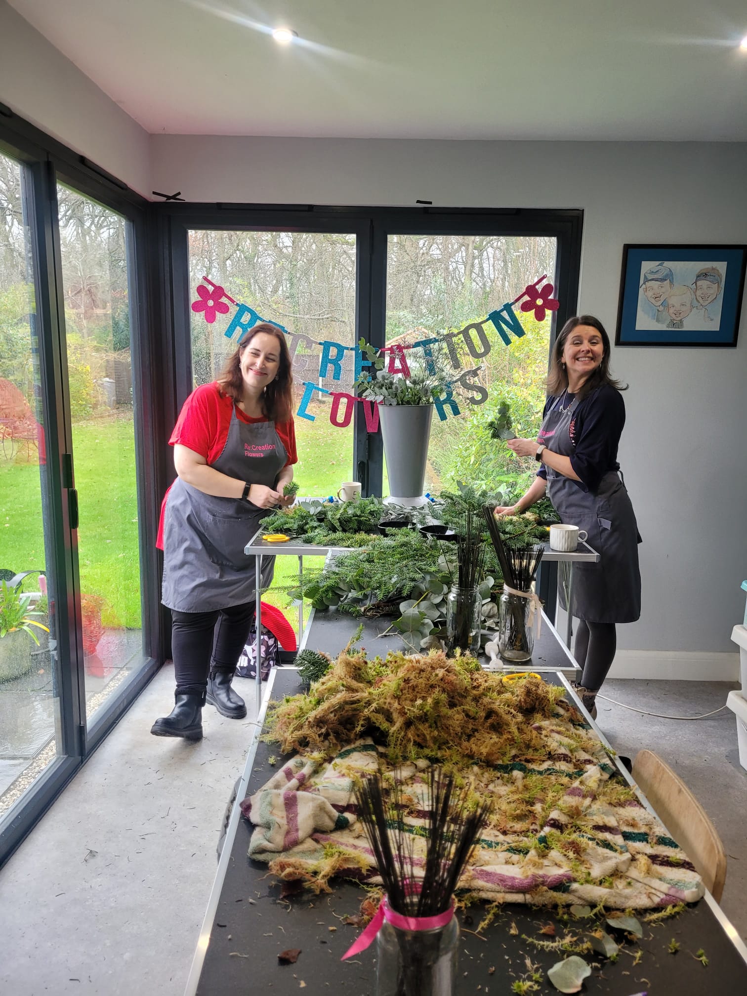 Two women in aprons stand by tables filled with festive greenery and moss, smiling warmly. Behind them, a colorful banner reads “Christmas Wreath Class” by ReCreation Flowers. Large windows reveal a lush garden outside.