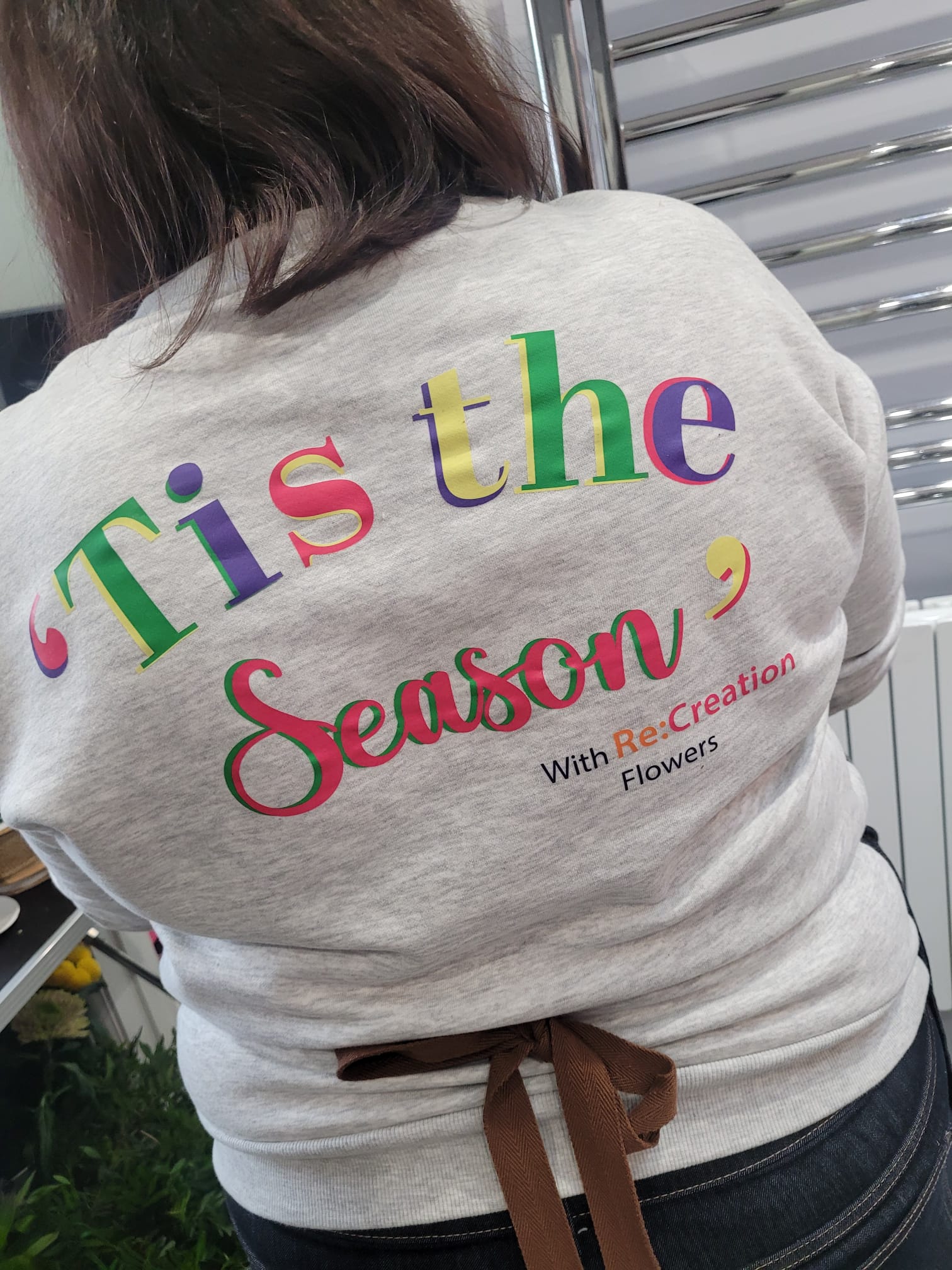 A person wearing a light gray sweatshirt with “Tis the Season” and “ReCreation Flowers” written in colorful letters on the back, standing indoors at a Christmas Wreath Class.