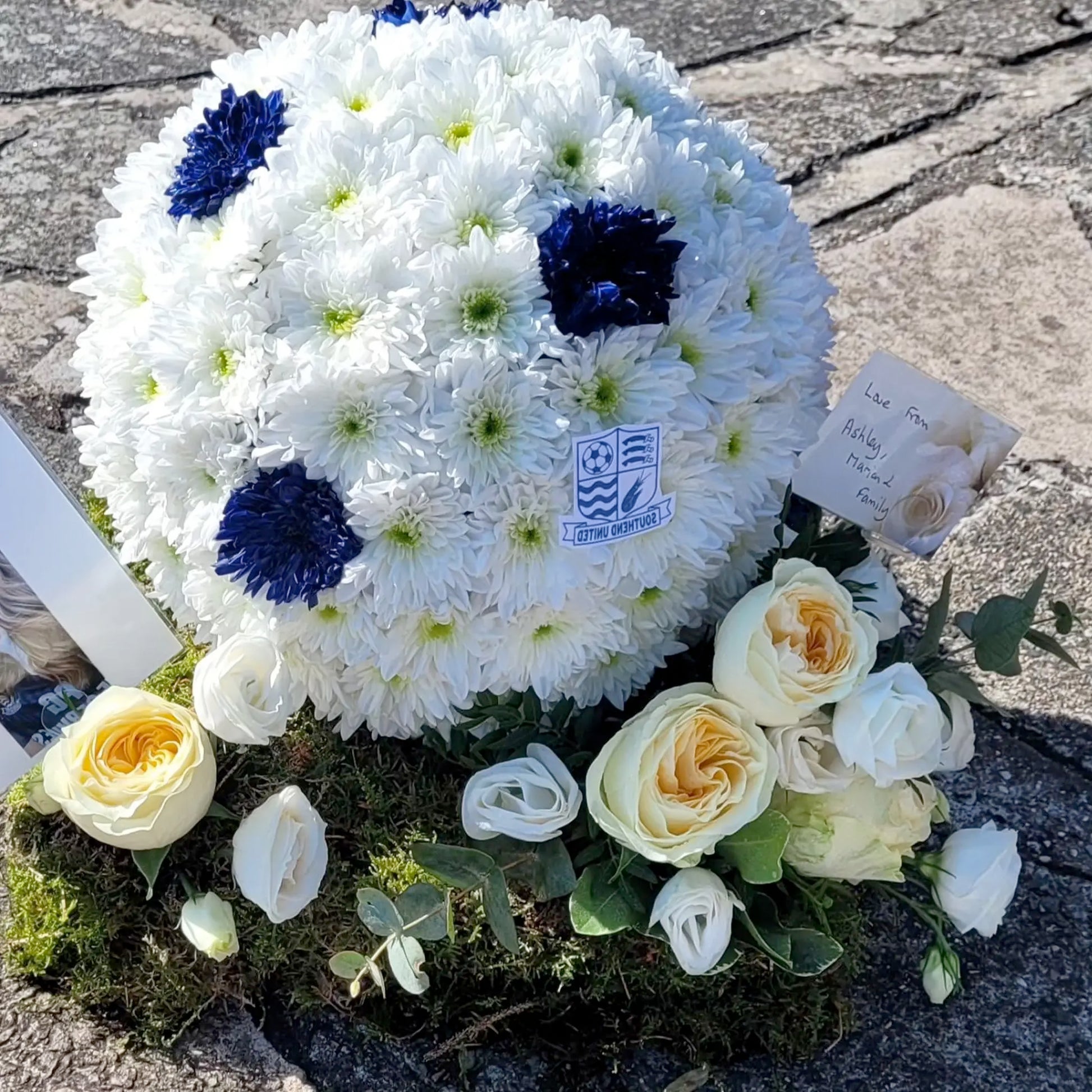 A soccer ball-shaped floral arrangement with white and blue flowers, Southend United badge, and card sits among roses—a perfect example of Special Tributes by ReCreation Flowers.