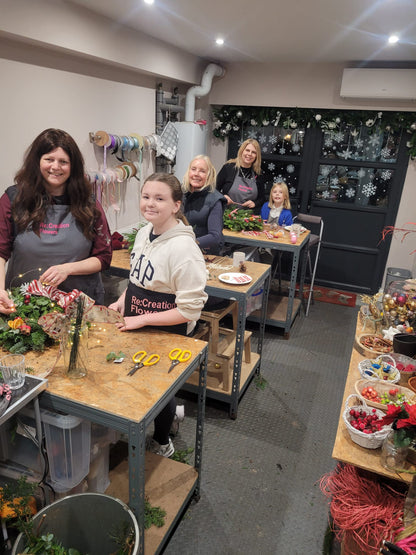 Five people, including two children, smile as they create festive floral arrangements during the Christmas Wreath Class by ReCreation Flowers. They work at stations in a craft room adorned with holiday garlands and snowflakes on the windows, with supplies and flowers scattered across the tables.