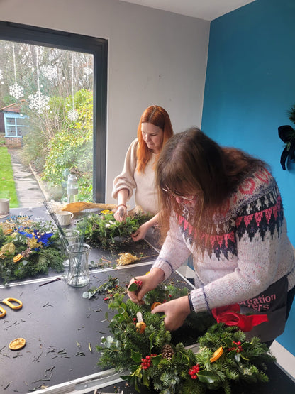 Two women participate in the ReCreation Flowers Christmas Wreath Class, crafting festive wreaths with greenery, pinecones, and berries at a table near a large window on a gray day. The blue-walled room overlooks a garden visible outside.