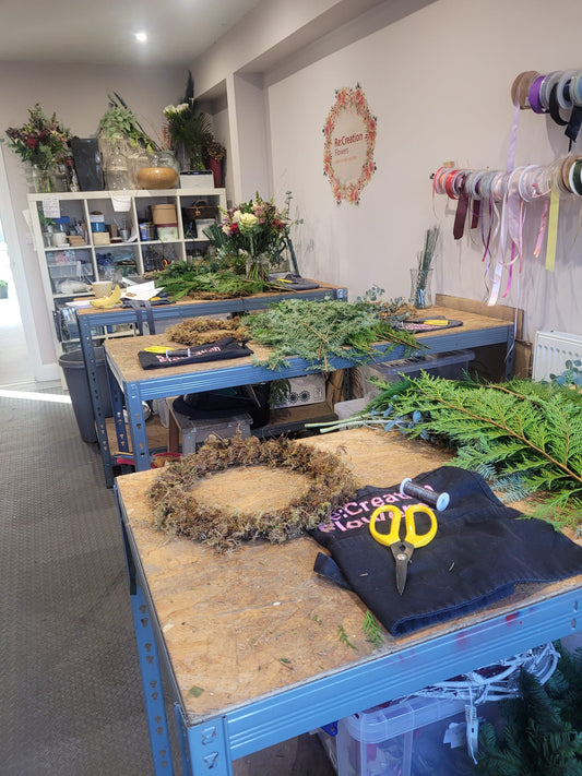 In the ReCreation Flowers studio, the Christmas Wreath Class features tables adorned with fresh greenery, wreaths-in-progress, and crafting tools like scissors. Shelves at the back are lined with vases and vibrant flowers, while a wall showcases colorful rolls of ribbon and a festive decorative sign.