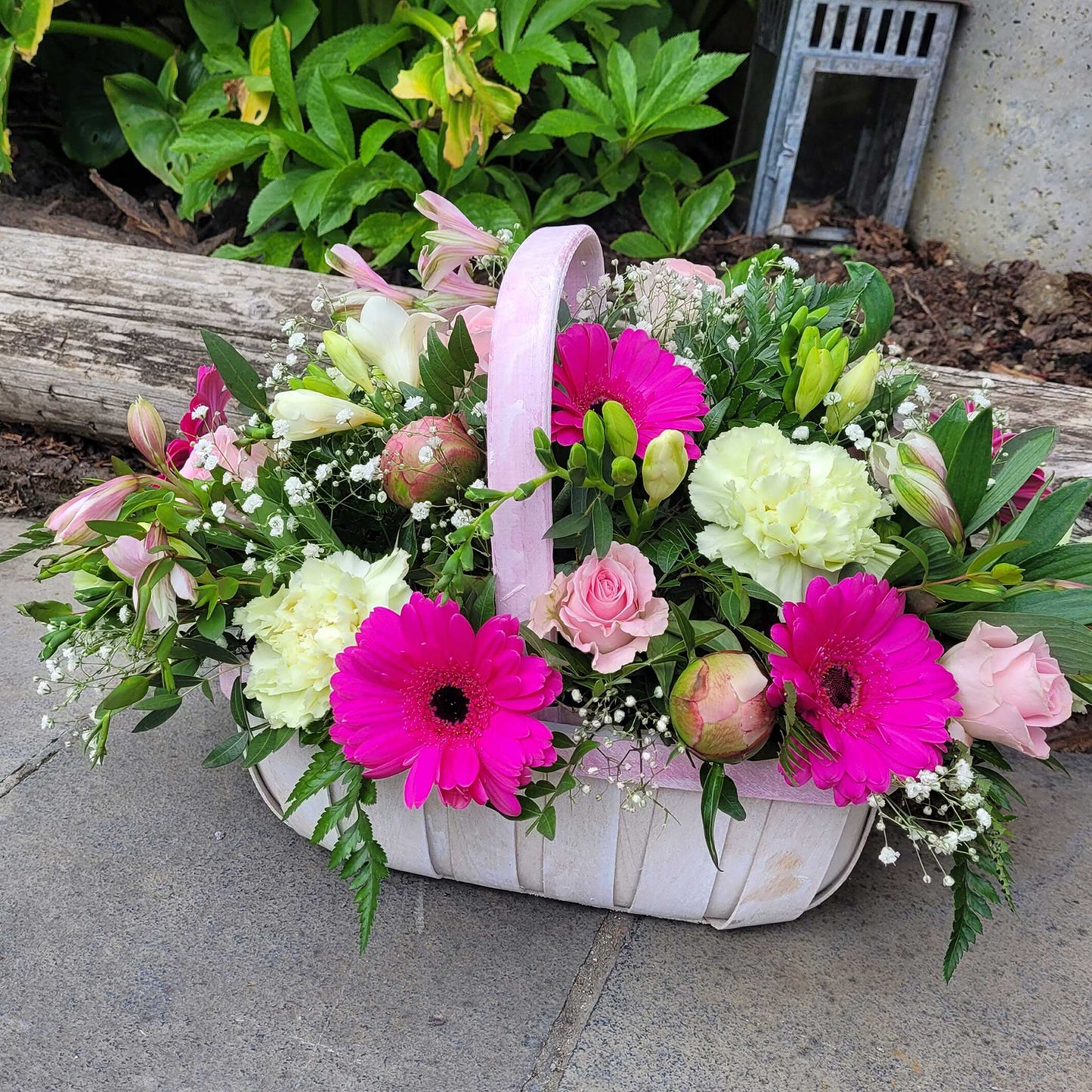 The Flower Arrangement by ReCreation Flowers features bright pink gerbera daisies, pale yellow carnations, light pink roses, greenery, and baby's breath in a white basket, displayed on a stone patio beside a log and lush green plants.