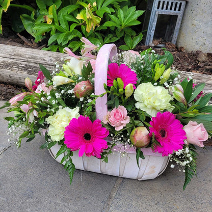 The Flower Arrangement by ReCreation Flowers features bright pink gerbera daisies, pale yellow carnations, light pink roses, greenery, and baby's breath in a white basket, displayed on a stone patio beside a log and lush green plants.