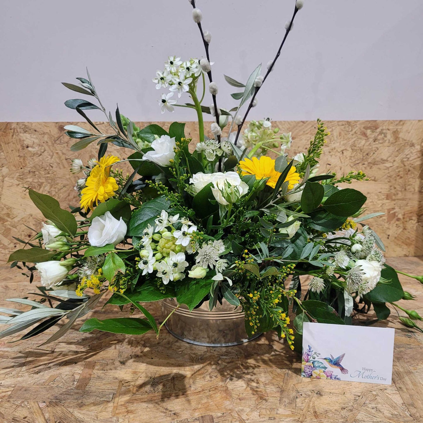 The Flower Arrangement by ReCreation Flowers features white and yellow spring blooms, green foliage, and pussy willow stems in a glass vase, with a hummingbird "Happy Mother’s Day" card displayed in front.
