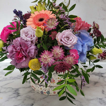 The Flower Arrangement by ReCreation Flowers features pink, purple, blue, and yellow spring blooms with lush green foliage in a decorative pot, set on marble—perfect for celebrations with hand-delivered flowers.