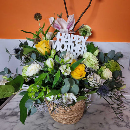 The Flower Arrangement by ReCreation Flowers features yellow roses, white blooms, pink lilies, and greenery in a woven basket with a Happy Mother sign. The orange and white marble background adds a stylish touch.