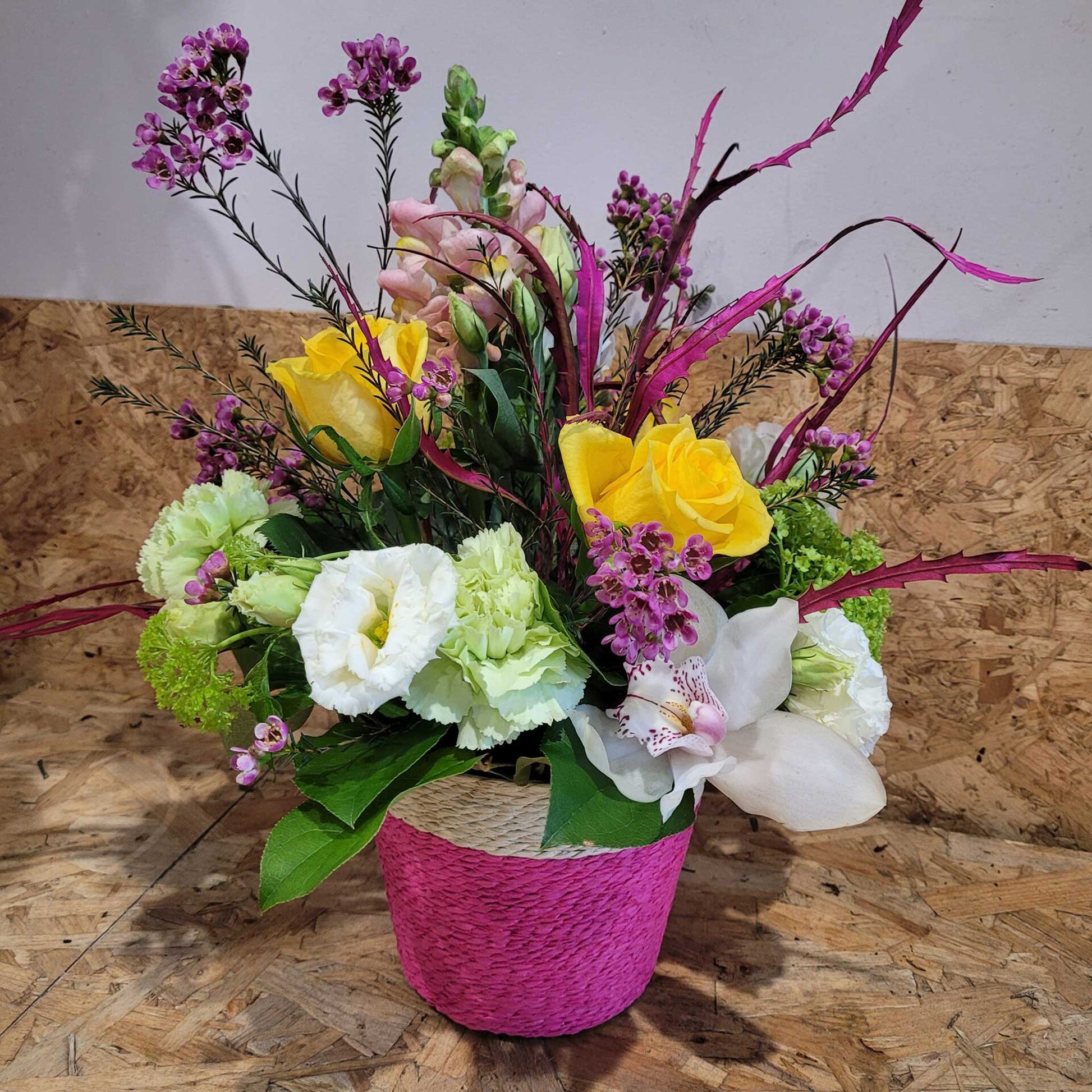 The Flower Arrangement by ReCreation Flowers features yellow roses, white lilies, spring blooms, and light green carnations in a pink basket—beautifully hand-delivered for any occasion.