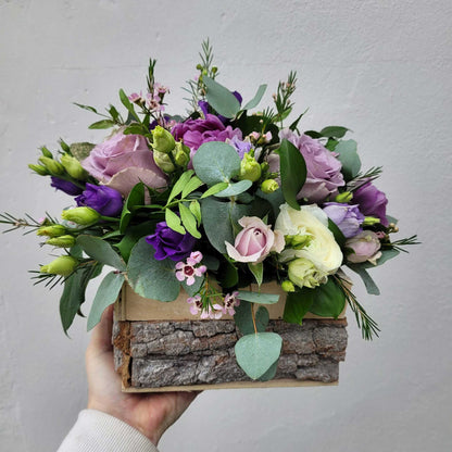 A hand holds the Flower Arrangement by ReCreation Flowers—a rustic box with bark, filled with seasonal purple, white, and pink blooms, green leaves, and eucalyptus—set against a plain light gray background.