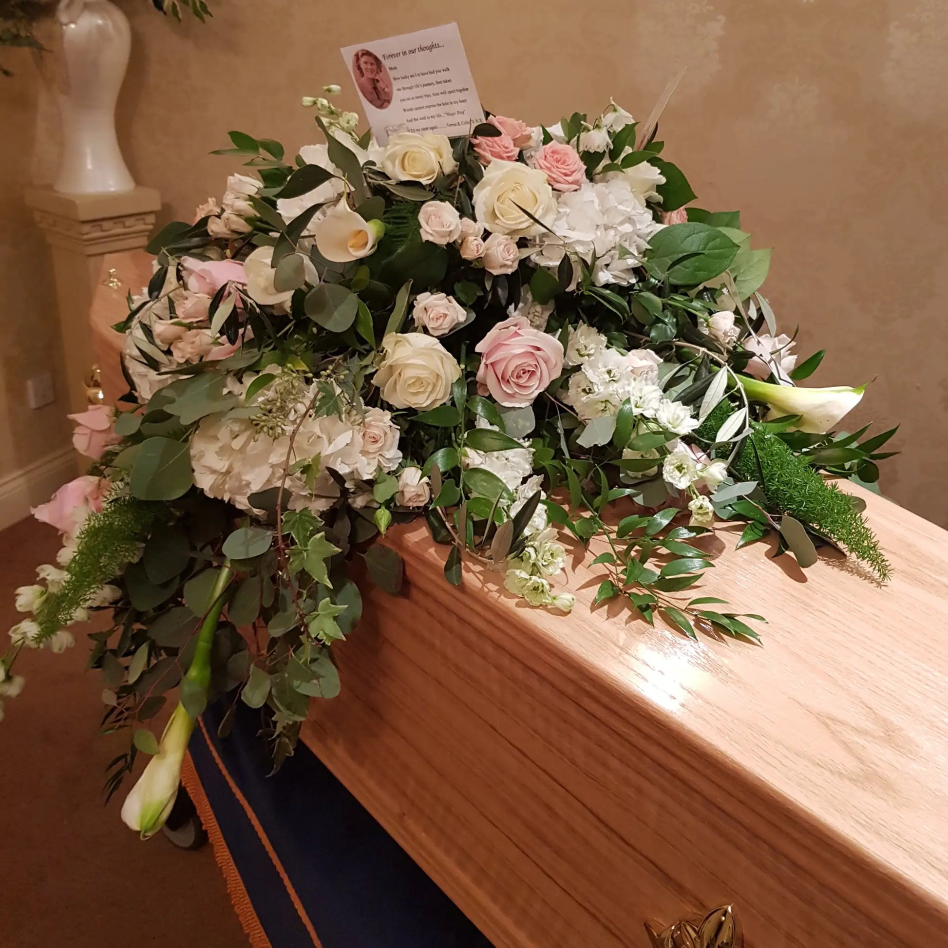 A wooden coffin is topped with ReCreation Flowers’ Casket Spray Funeral Flowers—lush white and pale pink blooms with greenery—and a card featuring a photo and message, set indoors for a celebration of life service.