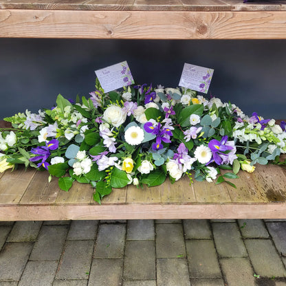 The Casket Spray Funeral Flowers by ReCreation Flowers features white and purple blooms, including roses and irises, arranged above a stone surface with two condolence cards nestled among the stems in a heartfelt tribute.