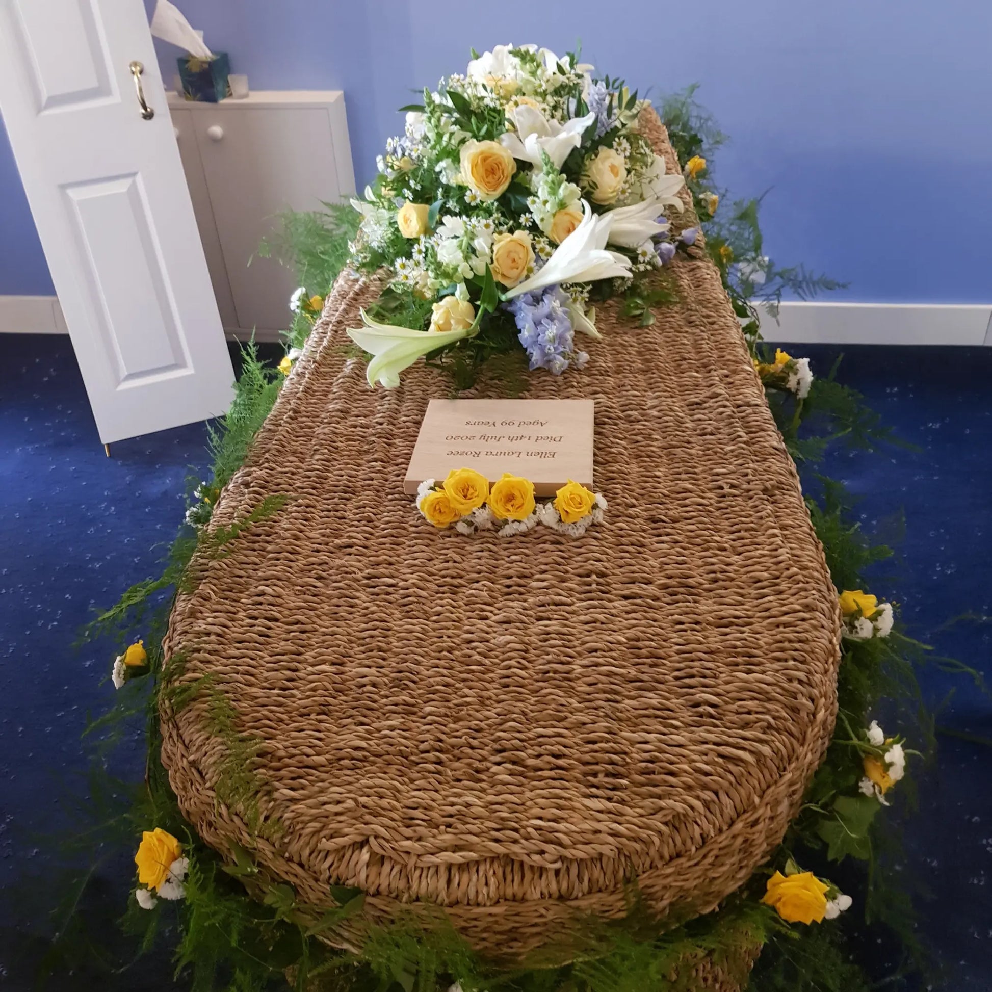 A wicker coffin with a Casket Spray Funeral Flowers arrangement by ReCreation Flowers, featuring white and yellow blooms with greenery, is set in a blue room. Yellow roses and a card rest on top, with an open door and cabinet in the background.