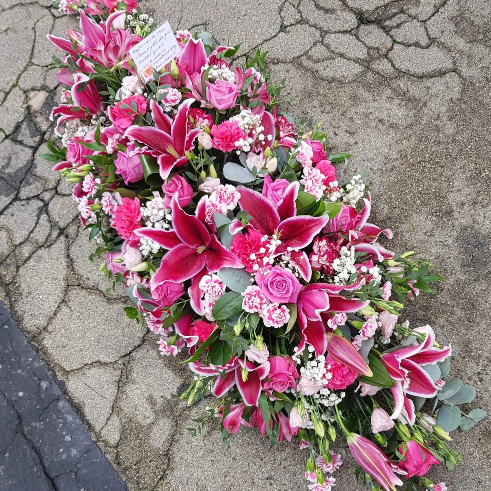 The ReCreation Flowers Casket Spray Funeral Flowers, featuring pink lilies, roses, and carnations, rests on cracked pavement. This elegant tribute, arranged with care and a small white card, honors a life celebrated.