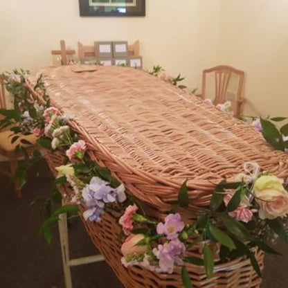 A wicker coffin with a ReCreation Flowers Casket Spray Funeral Flowers arrangement rests in a softly lit room with wooden chairs and a framed picture on the wall.