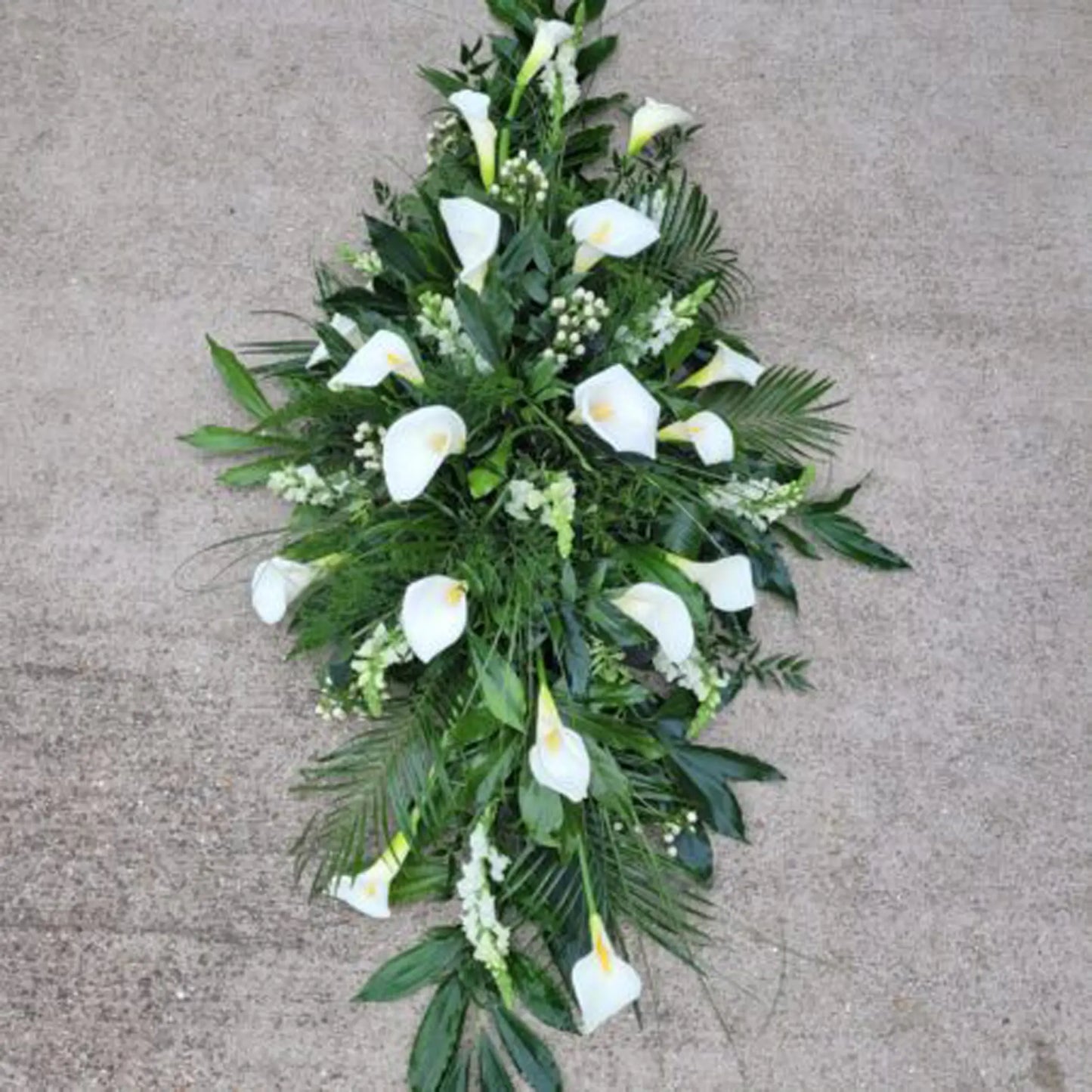 The Casket Spray Funeral Flowers by ReCreation Flowers features elegant white calla lilies and greenery, artfully arranged on a light concrete surface for a timeless tribute.