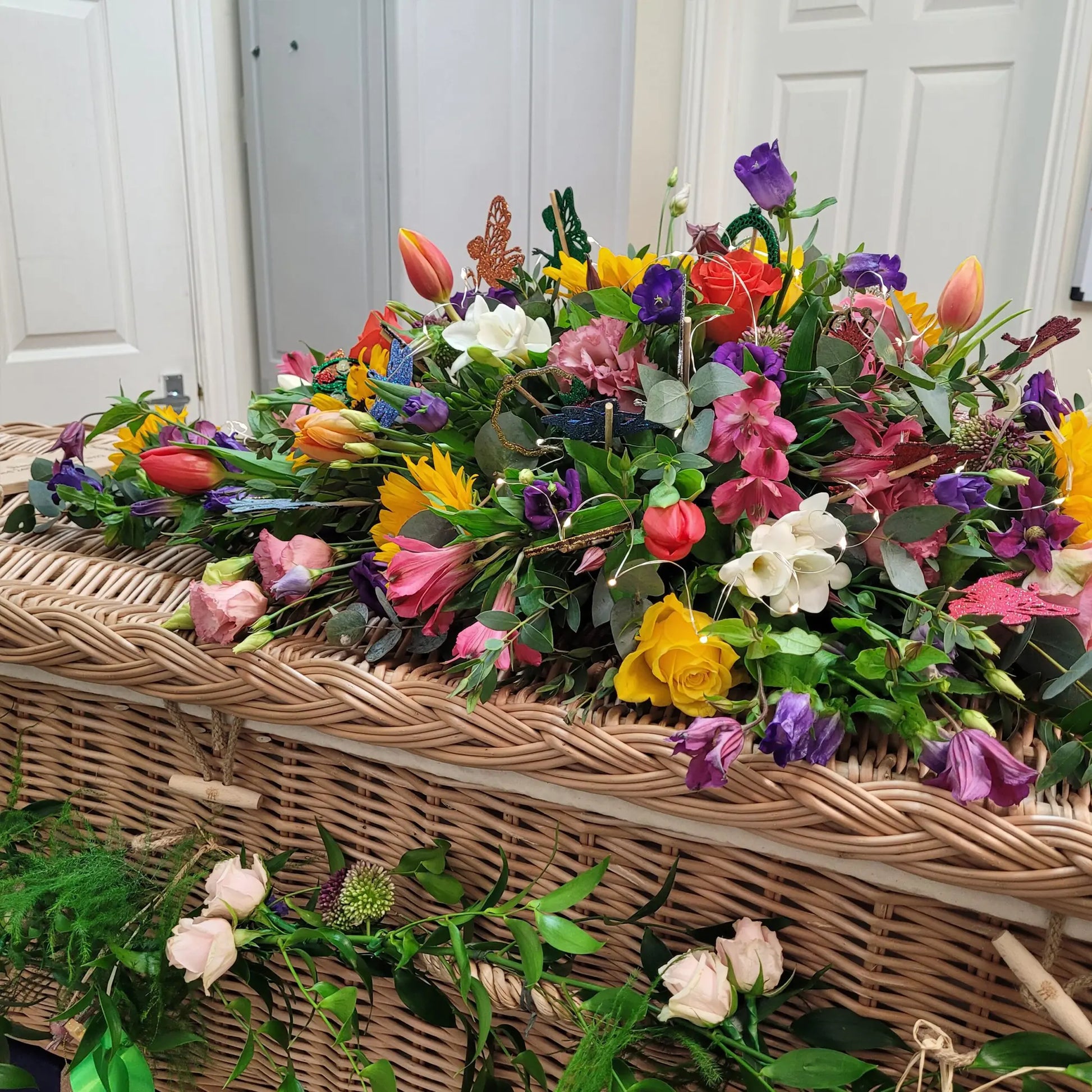 A wicker coffin with a Casket Spray Funeral Flowers arrangement by ReCreation Flowers—featuring sunflowers, roses, tulips, and greenery—stands in a room with white doors, honoring a celebration of life.