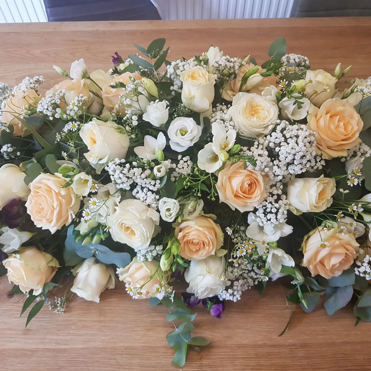 The Casket Spray Funeral Flowers by ReCreation Flowers features cream and pale peach roses, white freesias, baby's breath, and greenery—a tasteful tribute for a celebration of life.