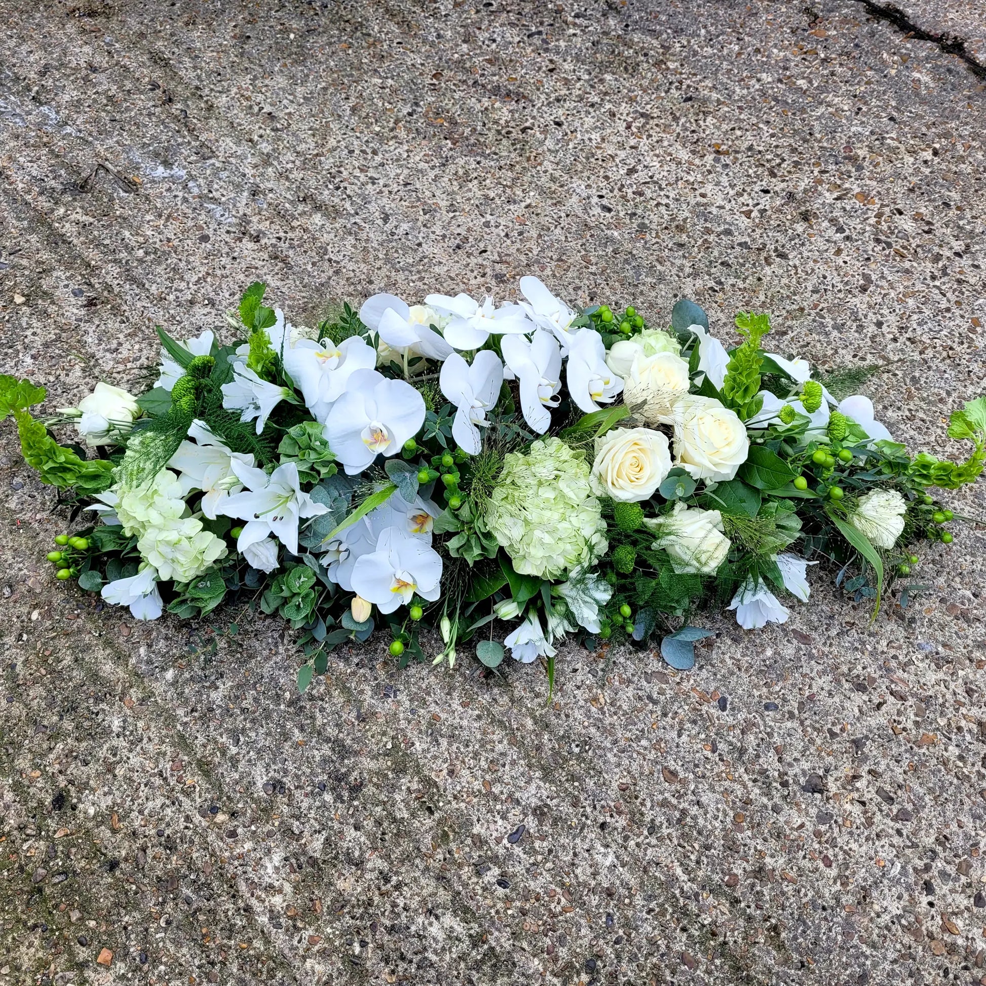 The Casket Spray Funeral Flowers by ReCreation Flowers features an elegant arrangement of white roses, orchids, and greenery displayed on a textured concrete surface.