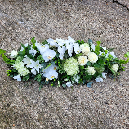 The Casket Spray Funeral Flowers by ReCreation Flowers features an elegant arrangement of white roses, orchids, and greenery displayed on a textured concrete surface.