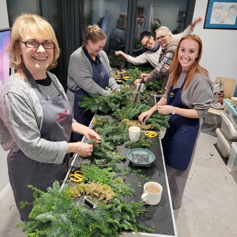 Five women smile and arrange greenery on a long table indoors, making crafts. Scissors, mugs, and other tools are on the table. The group wears aprons and casual clothes, and the atmosphere is cheerful and creative.