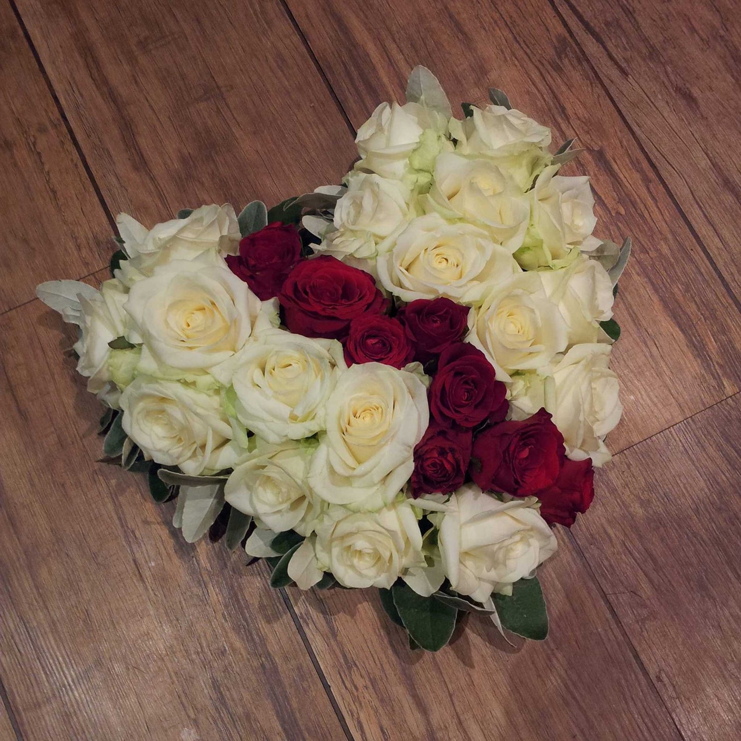 The Solid Heart Funeral Flowers - Loose Style by ReCreation Flowers features white roses outlining a heart and red roses clustered at the center—an elegant tribute, beautifully arranged on a wooden floor.