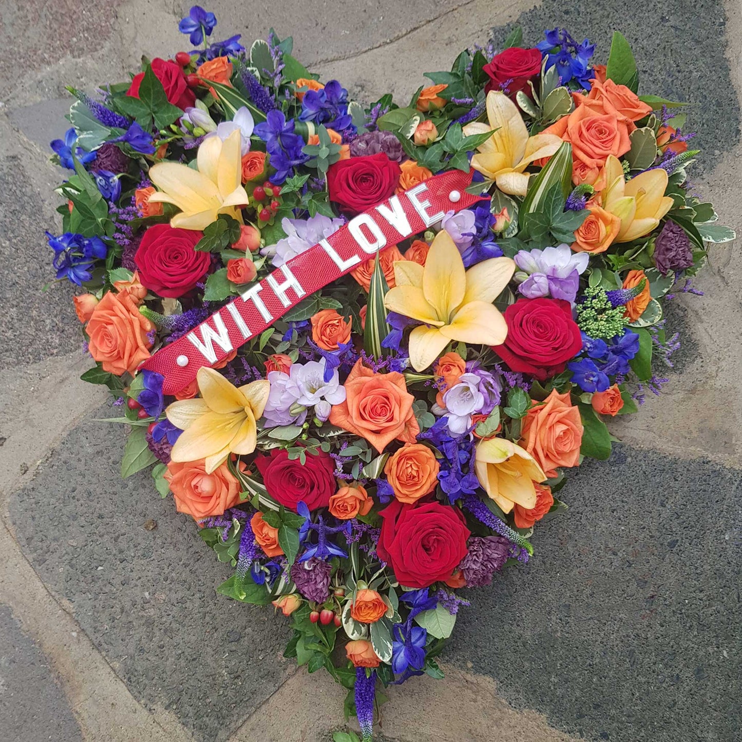 The Solid Heart Funeral Flowers - Loose Style by ReCreation Flowers features seasonal blooms in vibrant hues with a "WITH LOVE" ribbon, arranged on stone—an ideal heartfelt tribute for funerals.