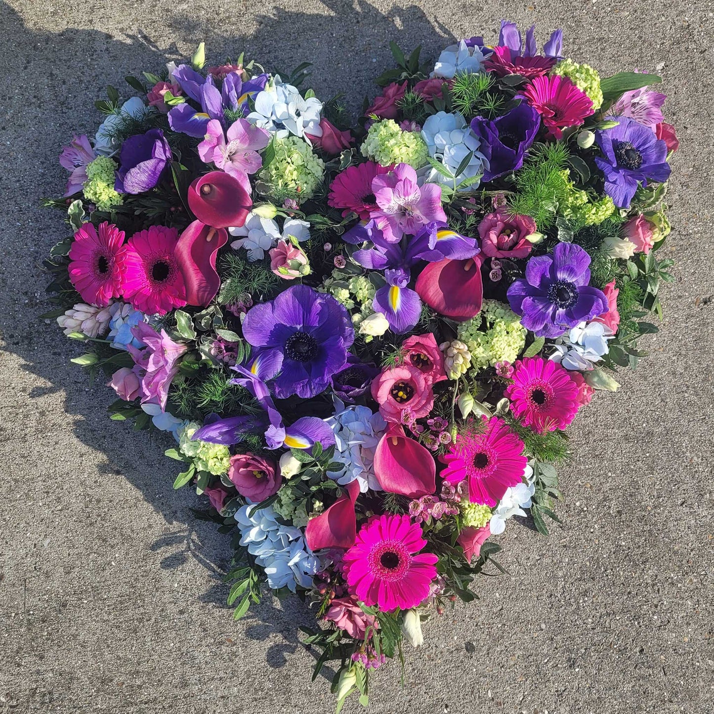 The Solid Heart Funeral Flowers - Loose Style by ReCreation Flowers features vibrant pink, purple, blue, and white seasonal blooms arranged in a heart shape—a stylish and elegant tribute for funerals.