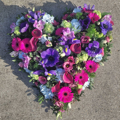 The Solid Heart Funeral Flowers - Loose Style by ReCreation Flowers features vibrant pink, purple, blue, and white seasonal blooms arranged in a heart shape—a stylish and elegant tribute for funerals.