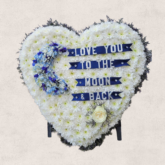 The Solid Heart Funeral Flowers by ReCreation Flowers is a heart-shaped arrangement of white blooms with a blue flower crescent, a white rose, and "LOVE YOU TO THE MOON & BACK" on blue ribbons.