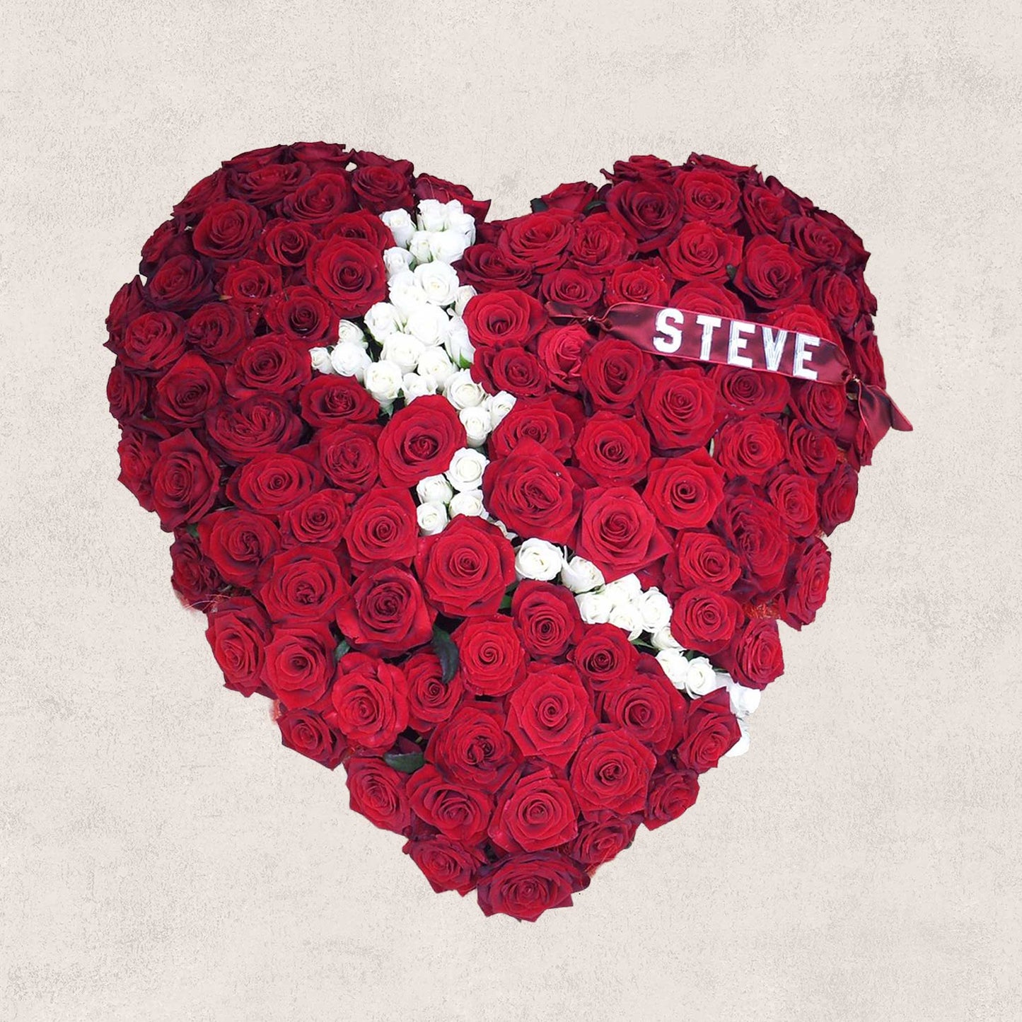 The Solid Heart Funeral Flowers - Loose Style by ReCreation Flowers is a red heart-shaped arrangement with a white rose cross at the center and a ribbon reading "Steve" on the upper right, set against a plain, light-colored background.