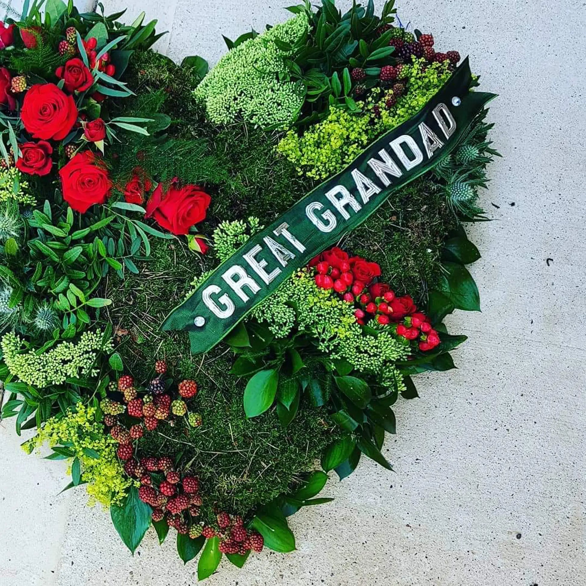 The Solid Heart Funeral Flowers - Loose Style by ReCreation Flowers features greenery, red roses, small red berries, and a "Great Grandad" ribbon, arranged on a light surface for a heartfelt tribute.