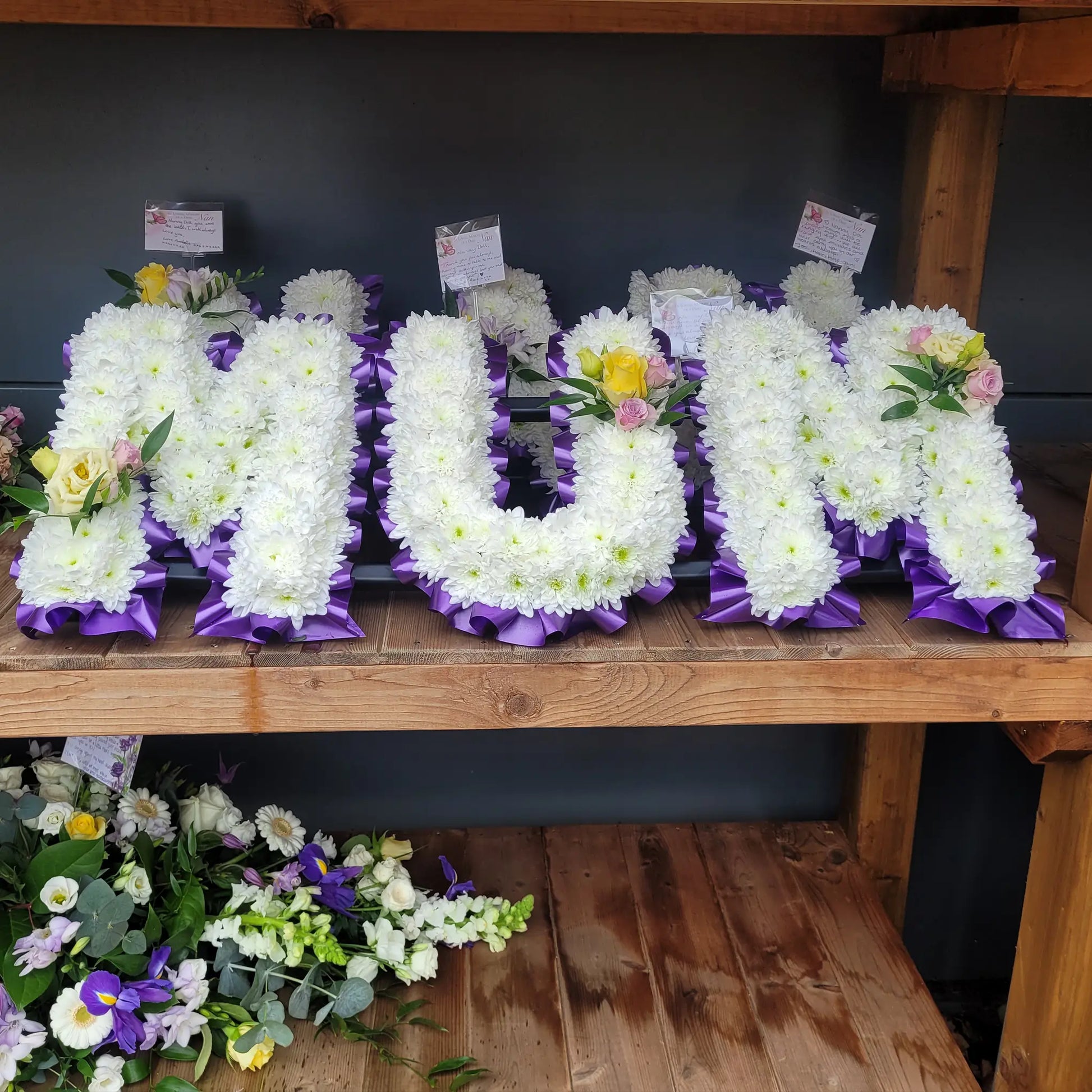 The Funeral Flower Letters – White Base by ReCreation Flowers feature "MUM" in large white blooms with purple ribbon edges, accented by yellow and pink flowers, creating a stunning custom funeral arrangement on a wooden shelf.