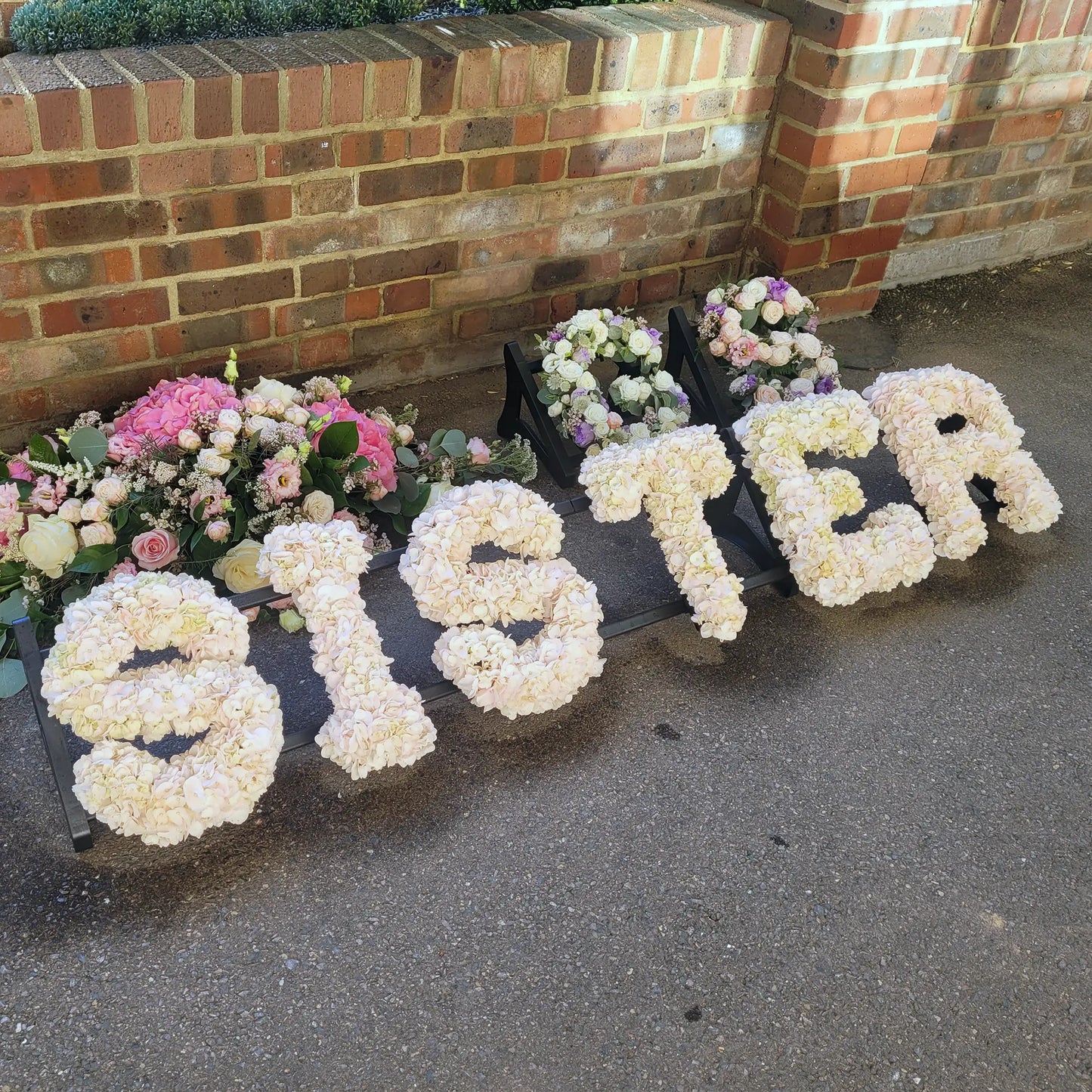 ReCreation Flowers’ Funeral Flower Letters – White Base spelling SISTER are arranged on the ground before a brick wall, surrounded by pink and white funeral arrangements.
