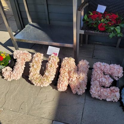 Funeral Flower Letters – White Base by ReCreation Flowers: large custom floral letters spelling "JUNE" in pale pink flowers, set on stone pavement beside red blooms and a card—a stunning massed display for any tribute.
