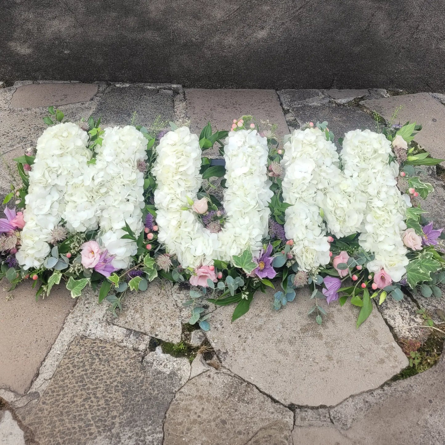 ReCreation Flowers’ Funeral Flower Letters – White Base spells "MUM" with white blooms, accented by greenery and pastel flowers, creating a heartfelt funeral tribute arrangement.