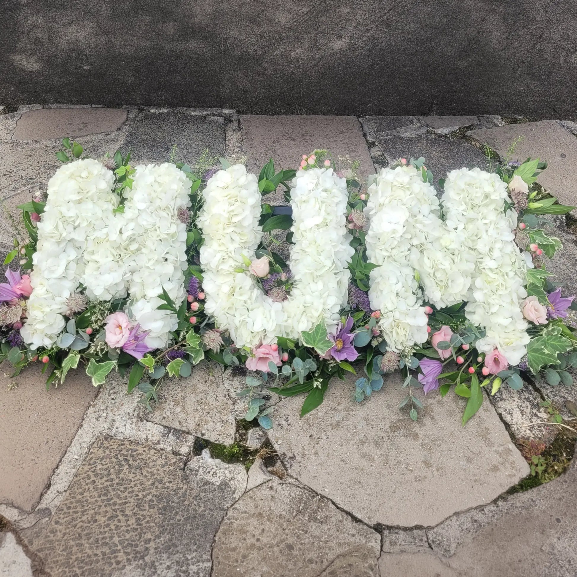 ReCreation Flowers’ Funeral Flower Letters – White Base spells "MUM" with white blooms, accented by greenery and pastel flowers, creating a heartfelt funeral tribute arrangement.