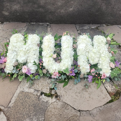 ReCreation Flowers’ Funeral Flower Letters – White Base spells "MUM" with white blooms, accented by greenery and pastel flowers, creating a heartfelt funeral tribute arrangement.