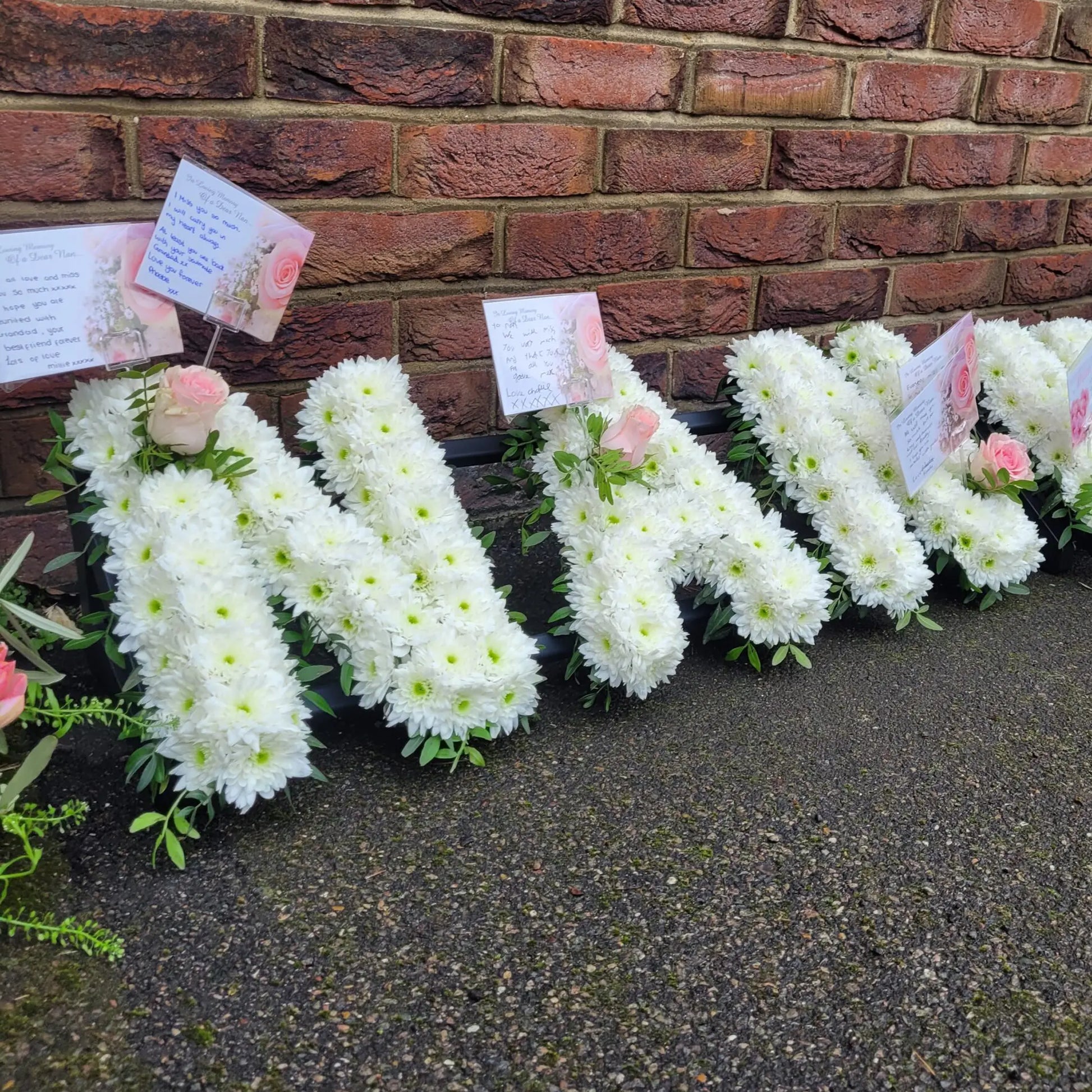 ReCreation Flowers' Funeral Flower Letters – White Base spell "MAM" against a brick wall, adorned with pink roses and message cards—creating a personalized tribute or memorial floral arrangement.