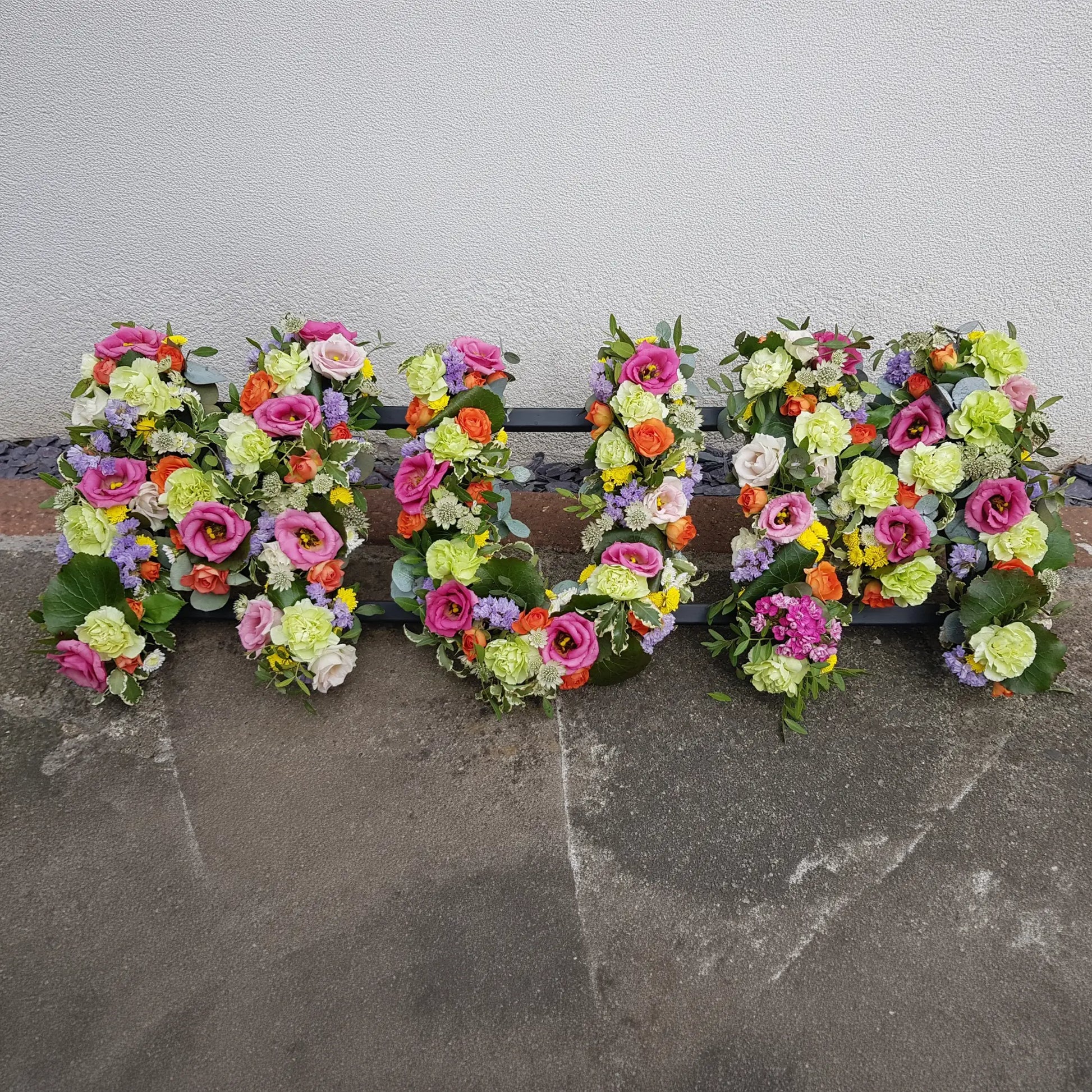 Funeral Flower Letters – Open Loose Style by ReCreation Flowers: large “MUM” letters crafted from fresh pink, green, orange, and white flowers, displayed against a light wall on a concrete surface.